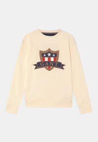 Krémová mikina s velkým emblémem, který zobrazuje štítový design v námořnické modré, červené a bílé barvě, s textem "GANT" na banneru.