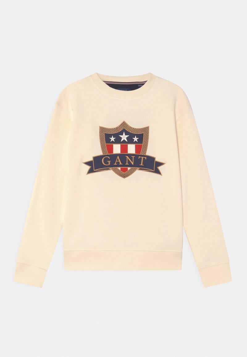 Krémová mikina s velkým emblémem, který zobrazuje štítový design v námořnické modré, červené a bílé barvě, s textem "GANT" na banneru.