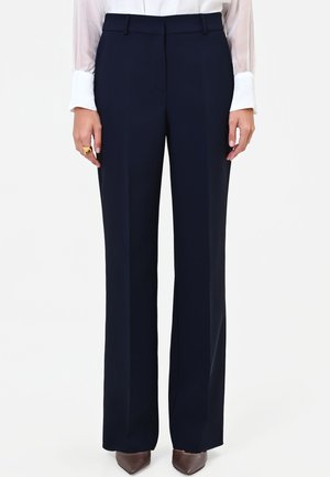 Donna che indossa pantaloni larghi blu scuro, camicetta bianca con maniche trasparenti, scarpe marroni a punta e un anello d'oro sulla mano destra.