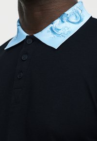 Κοντινή λήψη μαύρου πόλο πουκαμίσου με τρία μαύρα κουμπιά και ανοιχτό μπλε γιακά με σπειροειδείς κυματιστές σχεδιαστικές λεπτομέρειες.