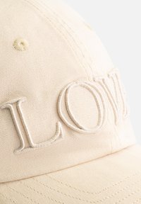 Beige baseballpet van geweven stof, met verhoogde "LOVE" borduring aan de voorkant en zichtbare stiksels langs de rand.