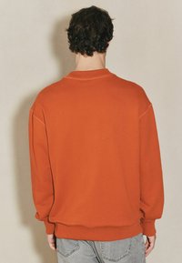 Pomarańczowy sweter z luźnym krojem, ściągaczami przy rękawach i dekolcie oraz prostymi detalami haftu. Nosi się go na jasnogranatowe dżinsy.