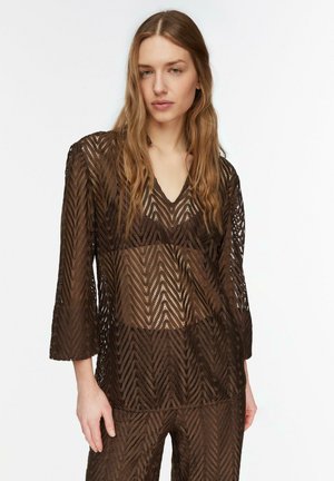 Młoda kobieta o długich blond włosach, ubrana w brązową prześwitującą koronkową bluzkę z dekoltem w serek oraz dopasowane spodnie z wzorem w jodełkę.