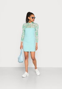 Vestido mini turquesa com mangas longas e transparentes com padrões; combinado com sapatilhas brancas e uma bolsa azul clara; modelo a usar óculos de sol.