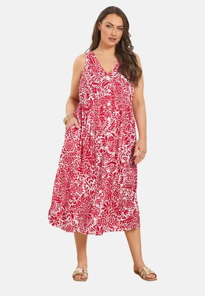 Kobieta w czerwono-białej sukience midi w kwiaty bez rękawów, z guzikami i kieszeniami, w beżowych sandałach i z bransoletkami.