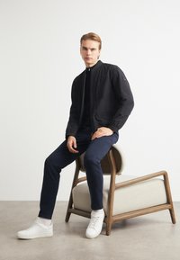 Black bomber jacket with ribbed collar, paired with navy slim-fit pants and white sneakers. Model seated on a wooden chair, light background.