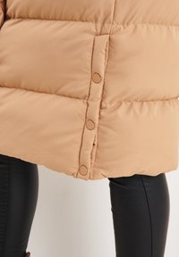 Beige puffer jacket with a quilted texture and round buttons along the hem, paired with black leggings. Smooth fabric with no patterns.