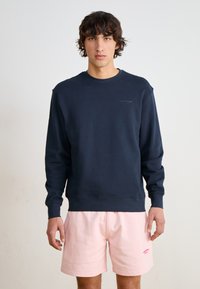 Sudadera azul marino con cuello redondo y puños de canalé, combinada con pantalones cortos de color rosa claro que presentan un logotipo. Tela suave, diseño casual.