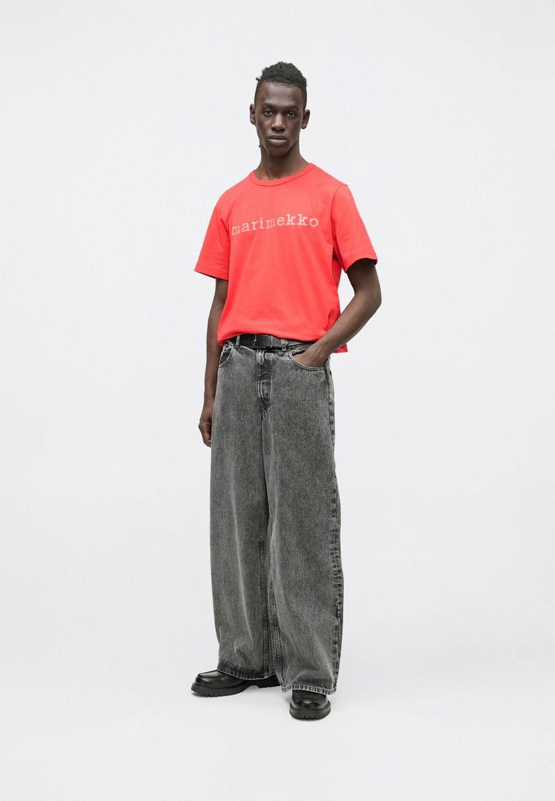 Czerwona koszulka z krótkim rękawem z napisem „marimekko” w jasnoszarym kolorze, w połączeniu z luźnymi, szarymi jeansami i czarnym obuwiem.