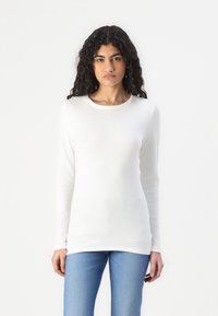 T-shirt bianca a maniche lunghe realizzata in tessuto morbido, con un colletto rotondo e una silhouette aderente, abbinata a jeans azzurri chiari.