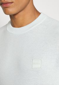 Hellblau strukturierter Pullover mit einem gerippten Rundhalsausschnitt und einem quadratischen weißen 'BOSS'-Logopatch auf der Brust.