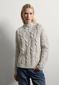 Un pull en tricot de câble gris clair avec un col montant, présentant un motif texturé et des poignets et un ourlet côtelés, porté sur un jean en denim bleu.