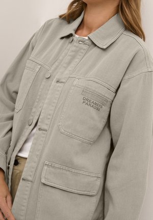 Personne portant une veste gris clair boutonnée avec de grandes poches, une poche brodée du texte « DREAMERS PARADISE ».