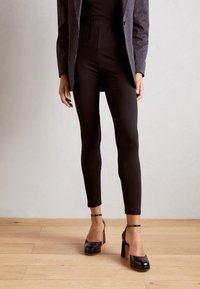 Leggings noirs en matériau extensible avec une texture lisse et longueur chevilles, associés à des talons en cuir verni noir épais.