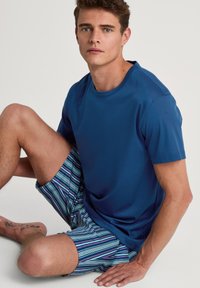 T-shirt blu a maniche corte abbinato a pantaloni corti a righe con linee blu, verdi e viola. Il tessuto appare morbido e leggero.