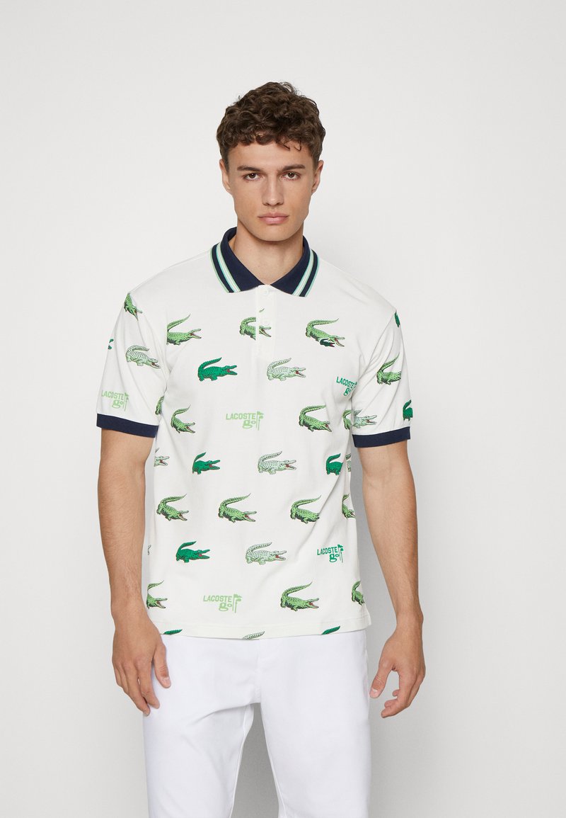 Polo blanc avec logos de crocodile verts et texte « LACOSTE ». Col marine avec rayures vertes. Pantalon en tissu blanc uni.