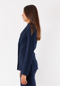 Blazer navy con design aderente, due tasche laterali e rever a punta. Il tessuto ha una texture liscia e una silhouette sagomata.