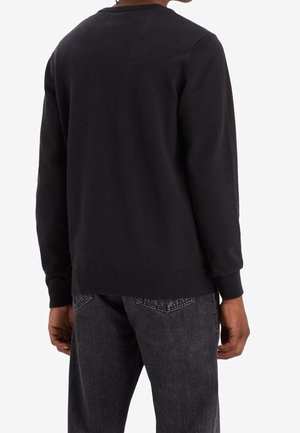Personne portant un sweat-shirt noir à manches longues et un jean gris foncé, vue de dos sur un fond blanc uni.