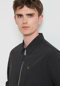 Junger Mann mit kurzen braunen Haaren, der eine schwarze zip-up Jacke mit Kragen und einem kleinen orangefarbenen Logo-Stickerei auf der Brusttasche trägt.