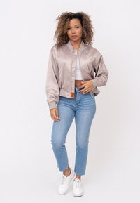 Satin bomberjack in lichtbeige met een slim-fit ontwerp, ribbelmanchetten en decoratieve patchdetails, gecombineerd met lichtblauwe jeans en witte sneakers.