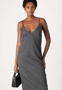 Grå dress med pinstriper, V-hals og tynne stropper. Har en tettsittende silhuett og glatt, teksturert stoff. Akkompagnert av en svart håndveske.