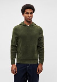 Maglione verde kaki con cappuccio, tessuto strutturato, tasca frontale e orlo a coste. Design strutturato con maniche aderenti, abbinato a jeans denim scuro.