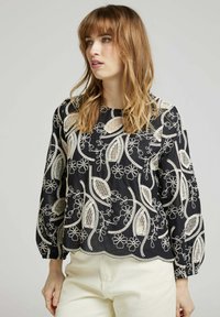 Blouse noire avec de délicates broderies florales blanches et des découpes. Elle possède un col rond et des manches longues élastiquées avec un ourlet festonné.