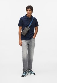 Jūras spēku teksturēta polo krekls, pelēki džinsi un rakstainā pleca soma ar zilo siksnu. Melni un zili sporta apavi pabeidz šo tērpu.