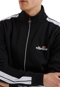 Svart zip-up jacka med hög krage, vita accentränder på ärmarna och en logotyp på bröstet, tillverkad av mjukt tyg.