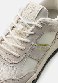 Sneaker beige e grigia con materiali in suede e mesh, cuciture tonali ben visibili, lacci e un sottile accento verde con branding.