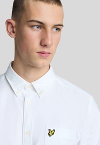 Camicia bianca con bottoni e taschino, caratterizzata da un logo giallo ricamato. La vista ravvicinata mette in evidenza il collo e la trama del tessuto.
