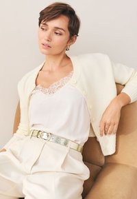 Kremowy kardigan na guziki, noszony na białą kamizelkę ozdobioną koronką oraz jasne spodnie, podkreślone metalicznym paskiem. Miękkie faktury.