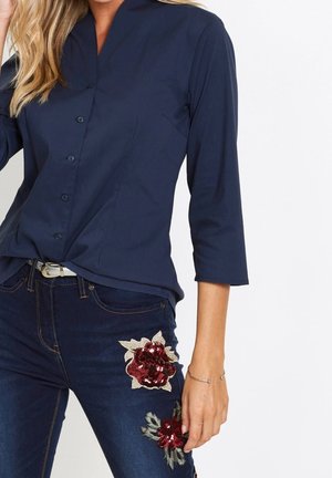 Femme portant une chemise boutonnée bleu foncé à manches trois quarts et un jean foncé avec des patchs de sequins rouges à motifs floraux sur la cuisse et le genou.
