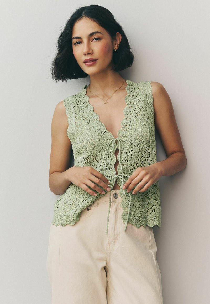 Gilet verde a maglia con bordi ondulati, motivo in pizzo e lacci frontali. Abbinato a pantaloni leggeri e larghi.