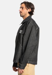 Black denim jacket with a pointed collar, front pocket, and branded patch. Features a loose fit and visible stitching details.