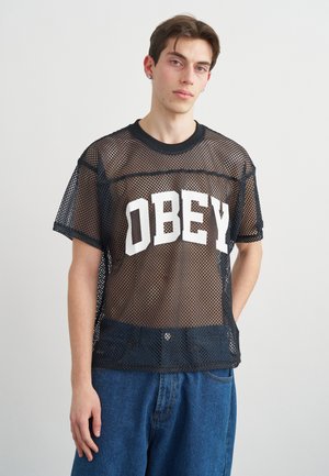 Νέος άντρας που φοράει μαύρο διαφανές T-shirt με μεγάλο λευκό κείμενο "OBEY" πάνω από μαύρο εσώρουχο και μπλε τζιν, στέκεται μπροστά σε απλό φόντο.