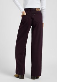 Dunkelviolette, weit geschnittene Jeans aus Baumwolle, mit einem hellbraunen Etikett am hinteren Bund und zwei Gesäßtaschen. Glatte Textur und taillierte Passform.