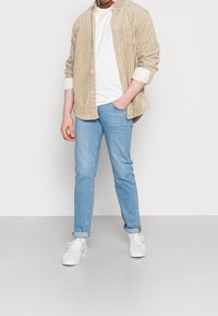 Béžová kordové košile přes bílém tričku, v kombinaci se světle modrými džíny a bílými teniskami. Rovný střih s vyhrnutými rukávy.