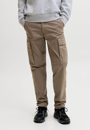 Personne portant un pantalon cargo beige avec des poches et des baskets noires, les mains dans les poches, debout devant un fond clair uni.