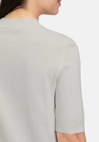 Jasnoszary sweter dzianinowy z krótkim kołnierzem i krótkimi rękawami. Posiada gładką fakturę oraz ściągacze przy mankietach i dolnym brzegu dla dodatkowego efektu.