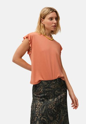 Mujer con una blusa coral de manga corta con volantes en las mangas, falda negra con estampado, collar amarillo y pendientes de aro dorados, mirando hacia la derecha.