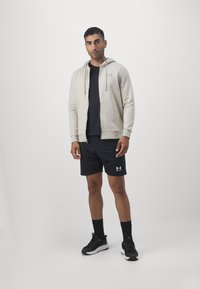 Uomo in piedi che indossa un felpa beige con zip, una maglietta nera, pantaloni corti neri, calze nere e scarpe da ginnastica nere su uno sfondo bianco semplice.