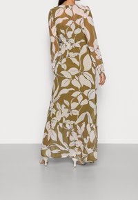 Pitkä, liehuva mekko oliivinvihreänä, jossa on valkoisia lehtikuvioita, pitkät hihat, kerätty vyötärö ja röyhelöhelma.