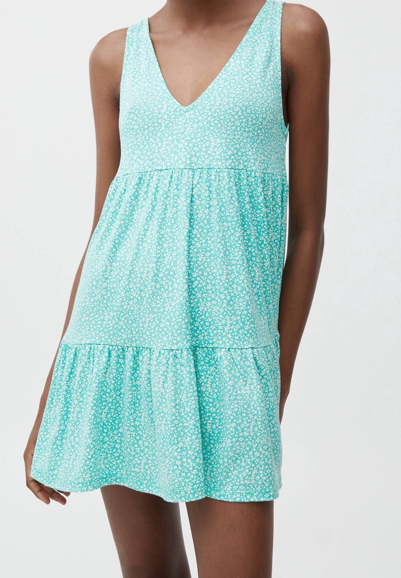 Robe turquoise clair avec un décolleté en V, sans manches, taille froncée et ourlet volanté. Présente un petit motif floral blanc sur l'ensemble.
