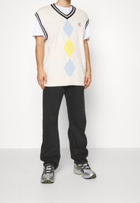 Mężczyzna w kremowym bezrękawniku w wzór argyle, noszący białą koszulkę, czarne spodnie i srebrne trampki z czarnymi i żółtymi akcentami.