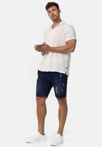 Polo majica kratkih rukava u teksturiranoj krem boji s gumbima, u kombinaciji s tamnim traper šortsevima s poderanim detaljima i horizontalnim šavovima. Bijeli tenisice.