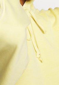 Světle žlutá mikina s kapucí vyrobená z měkké tkaniny, s šňůrkovým zapínáním a žebrovanou texturou. Obsahuje vyšívaný detail loga.