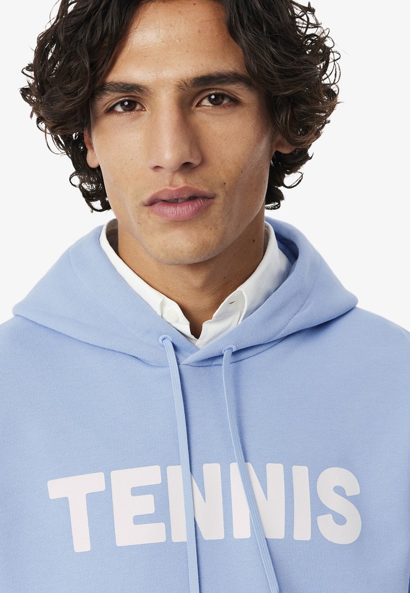 Giovane uomo con capelli ricci scuri che indossa una felpa con cappuccio azzurro chiaro con la scritta "TENNIS" sul petto e una camicia bianca con colletto sotto.