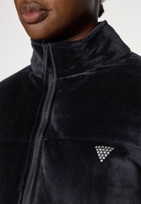 Giacca nera in velluto con zip, caratterizzata da un colletto alto e un triangolo decorativo con strass sul petto. Tessuto morbido al tatto.