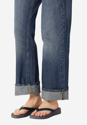 Blote voeten met donkerblauwe geweven slippers en opgerolde blauwe spijkerbroek tegen een effen achtergrond.